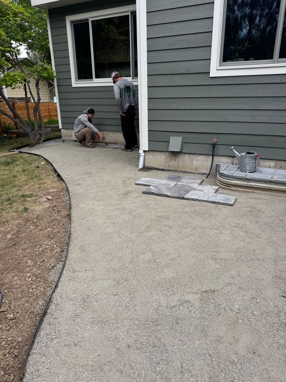Two workers preparing a gray gravel walkway beside a house with green siding.