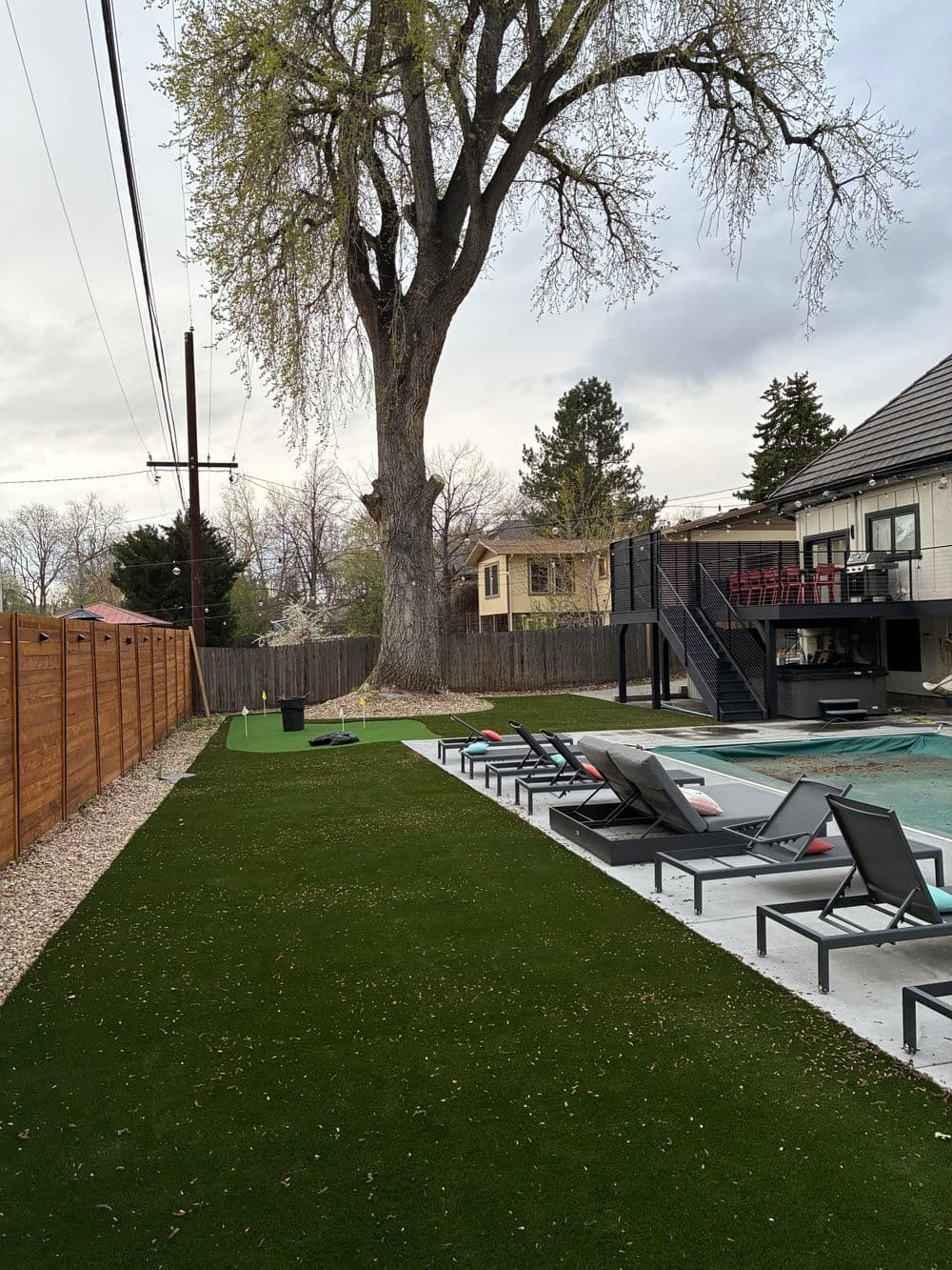Backyard view featuring a pool, lounge chairs, and a large tree under a cloudy sky.